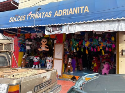 A vibrant piñata shop, "Dulces y Piñatas Adrianita," displays a colorful array of piñatas under a blue awning. The shop's entrance overflows with character piñatas: animals, cartoon figures, and traditional stars. A baby piñata hangs prominently. In front, a rusted pickup truck partially obscures the view. A vehicle's hood is visible in the foreground, suggesting the photo was taken from a car.