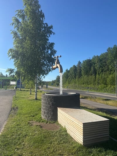 A bronze-colored faucet sculpture is positioned on a low, circular stone structure. Water flows from the faucet into a basin below. Beside the stone structure is a stack of light-colored wooden planks, likely forming a bench or seating area. A large tree with green leaves stands to the left of the sculpture, and several other trees and a dense forest border the background. A paved road with painted lines runs parallel to the forested area. On the far left, a green canopy structure, possibly a bus stop or fuel station shelter, is visible. The scene is illuminated by bright sunlight under a clear blue sky, indicating an outdoor, daytime setting. This artwork is located at a rest stop along the E75 highway in Heinola, Finland.
