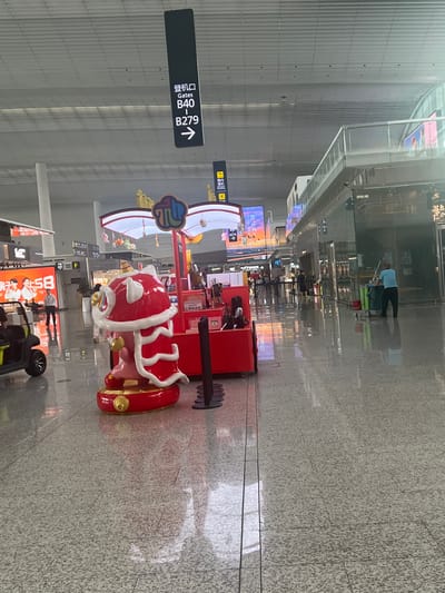 This is a photo of Guangzhou Baiyun International Airport in Guangzhou City, China. The image shows a wide, well-lit interior with a polished floor that reflects the overhead lighting and the surrounding architecture.
In the foreground, a decorative red and white lion statue, likely a Chinese New Year decoration, stands on a red base. Behind it is a red stall with a white canopy, adorned with small colorful hanging ornaments. To the left of the lion statue, a yellow golf cart-like vehicle is visible.
Further into the background, there are signs indicating "Gates B40 - B279" with an arrow pointing to the right, suggesting a direction within the airport. Several people are seen walking in the distance. Shops with glass fronts are visible along the right side of the image, and one person is operating a cleaning cart, mopping the floor. Large digital screens displaying graphics and text are also present in the background, adding to the modern airport environment. The overall atmosphere appears to be one of a busy but organized transportation hub.