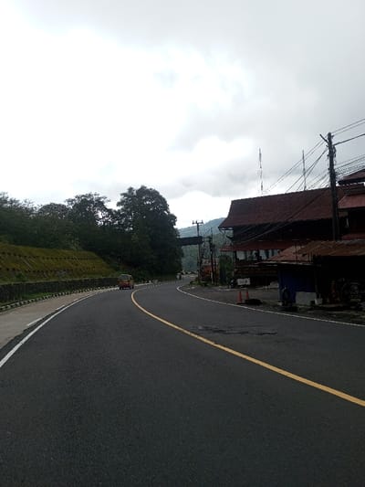 The scene captures a section of a curved, two-lane asphalt road in Deothang, Bhutan, under an overcast sky. A solid yellow centerline and a white left shoulder line delineate the road. A light-colored van, likely beige or light yellow, is observed traveling away from the viewer on the left curve of the road. To the immediate left, a manicured green terraced hillside is contained by a low concrete and stone retaining wall, transitioning into dense foliage further uphill. On the right side of the road, a cluster of buildings with reddish-brown, likely corrugated metal, roofs are present. The largest building features multi-tiered wooden structures. Utility poles with multiple electrical lines are positioned along the roadside on the right. A smaller blue-painted structure is visible at ground level, alongside two orange traffic cones. Distant forested hills are partially obscured by the heavy cloud cover.