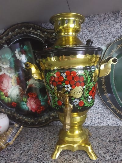 The image features a close-up, top-down view of a decorative golden samovar resting on a tray adorned with floral and bird motifs. The samovar itself is intricately decorated with a black base featuring a vibrant, hand-painted design of red berries, green leaves, and small white flowers. A brass spigot protrudes from its side. The tray beneath it is also black, elaborately painted with a profusion of colorful flowers, including pink roses and white daisies, and a small bird with white and grey plumage.
The samovar and tray are placed on a speckled grey surface, likely granite or marble, which suggests an indoor setting, possibly a home or a shop display. In the background, a portion of a dark green, circular object with a decorative gold trim is visible, hinting at another decorative item or surface. To the right of the samovar, a gold-colored handle or spout is clearly depicted, and a part of what appears to be a beaded necklace or decorative string is visible in the lower right corner.
There is no discernible activity taking place in the image; it is a still life arrangement. The lighting appears to be artificial, casting highlights on the polished surfaces of the samovar and tray. The overall impression is one of traditional artistry and decorative craftsmanship. No text is visible in the image. The location context of Waverley, United Kingdom, does not offer specific visual cues within the image itself.
