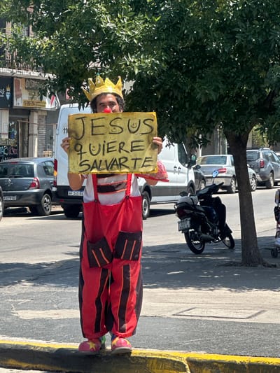 A person in a clown costume, complete with a red nose and a gold crown, stands on a city street holding a sign. The sign, written in Spanish, proclaims "Jesus quiere salvarte" or "Jesus wants to save you." The individual is positioned near a sidewalk curb and parked cars. A tree with dense foliage looms in the background. A motorcycle is also parked nearby.