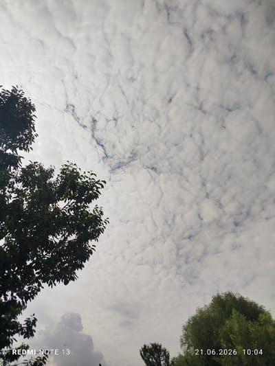 The image shows a cloudy sky with trees in the foreground and background. A plane is visible in the distance. The lower left corner of the image contains the text "REDMI NOTE 13", and the lower right corner contains the text "21.06.2026 10:04". The location is stated to be Асан-Чек, Kyrgyzstan. The clouds appear to be altocumulus. The trees are lush and green, suggesting a temperate climate and possibly the summer season. The overall lighting suggests it is daytime, likely morning or afternoon given the timestamp. There are no people or specific interactions depicted.