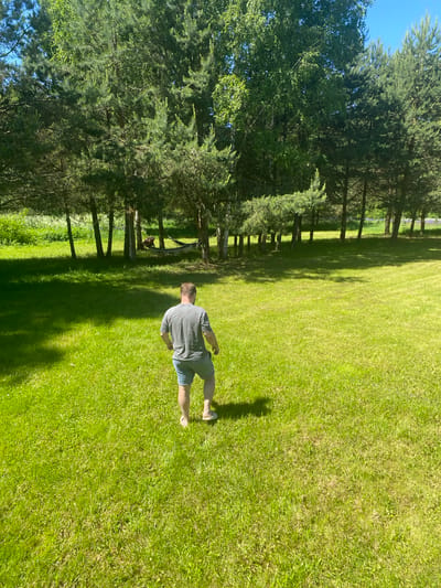 The scene, set in Копнино, Russia, depicts an outdoor environment on a sunny day. A male subject, viewed from behind, walks across a vast expanse of bright green grass towards a line of varied trees in the background. He wears a grey t-shirt, light blue denim shorts, and light-colored flip-flops. His body is oriented with his back to the viewer, and his left leg is mid-stride. Shadows from trees and the subject are cast upon the grass, indicating strong sunlight from the upper right.
The background features a dense line of trees, including several coniferous pine trees with dark green needles and multiple deciduous birch trees with lighter, often white, bark. Between two tree trunks on the left side of the background, a light-colored hammock is suspended, containing an indistinct figure or object, likely a person resting, largely obscured by shadow. Beyond the tree line, a lighter green field or clearing is visible, with subtle hints of purple vegetation, possibly flowers, further in the distance on the right side. The sky in the upper corners is clear and blue.