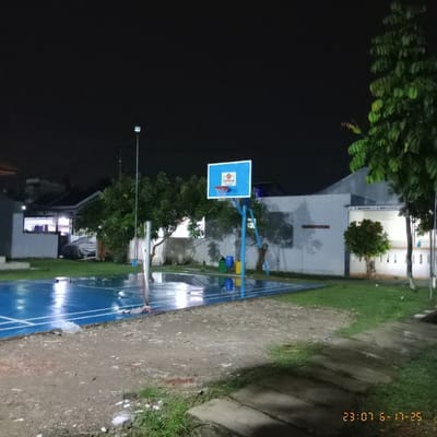 A low-angle, long shot presents a nighttime scene of a blue basketball court. A basketball hoop is visible in the center. The court surface is reflective, appearing wet. A building is behind the court. Trees flank the area. The sky is dark, suggesting a late hour. A sidewalk runs along one edge, with grass on the other side. The date "23 07 16-17-25" is displayed in the upper right corner.