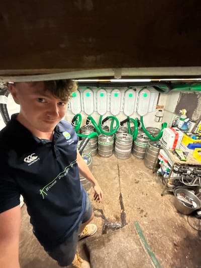 The image depicts a young Caucasian male taking a selfie within an interior space, identified as a beer cellar or utility room. The male subject, positioned in the lower-left foreground, has short, curly brown hair and is wearing a dark blue polo shirt with a Canterbury logo, a "Newfoundland Tour 2023" patch, and "A Home Design Concept" text. He also wears grey shorts and tan work boots, and is looking directly at the camera with a slight smile.

The background reveals a row of eight white, wall-mounted units, likely part of a beer line cleaning system, each displaying a green indicator light. Below these units, approximately ten to twelve silver-colored beer kegs, some bearing "TETLEY OF HEINEKEN" branding, are arranged on a concrete floor. Green flexible hoses connect the kegs to the wall-mounted units. The room features a low, unfinished ceiling, white-painted walls, and a concrete floor exhibiting stains, moisture, and a prominent crack. To the right, a makeshift shelf or workbench holds various items including a white box, yellow bins, a spray bottle, and other tools. No specific identifiers for the location in Sigglesthorne, United Kingdom, are visible within the image.