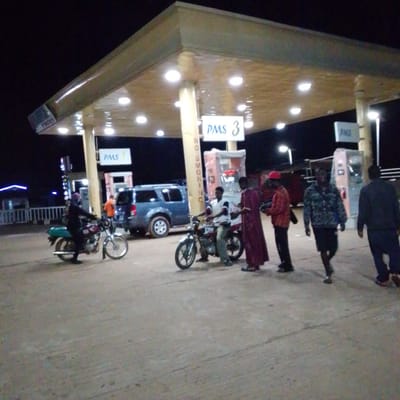 At night, a gas station in Mangu, Nigeria, is visible. The gas station has a covered canopy with lights illuminating the area. Several people are present, some interacting with motorcycles. One person is on a motorcycle, while others are standing or walking. A blue SUV is parked near a fuel pump. Signs for "PMS" and "NO SMOKING" are visible on the pillars supporting the canopy. The ground is paved, and the background is dark, indicating it is nighttime.