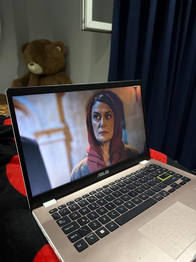 The image depicts an open laptop displaying a scene of a woman wearing a dark red hood with light reflecting on her face. The laptop is positioned on a surface with a red and black circular pattern. The keyboard of the laptop is visible, with black keys and some alphanumeric characters labeled. In the background, a blurred teddy bear sits on a grey surface, next to a window frame. The scene is indoors, with a dark blue curtain visible in the upper left corner of the image. There is no information regarding the location in Vinitsa, Bulgaria.