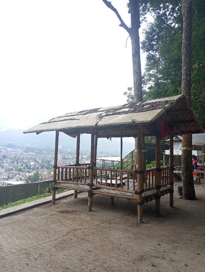 The scene is an elevated outdoor viewing platform in the city of Gadog, Indonesia. A vacant, open-sided gazebo, constructed from light-colored bamboo poles secured with dark lashings, occupies the center. Its pitched roof is covered with a sheet material overlaid with dried foliage or thatch, and a red fabric hangs from its interior right side. The gazebo features raised seating with pebble infills and a low central platform. To the right of the gazebo, two large tree trunks rise from the concrete ground, wrapped with yellow bands. Beyond these trees, a white tent-like structure and an additional outdoor seating area with tables are visible. Two individuals are present in this background area on the right; one is standing, wearing a pink top. The foreground consists of an empty concrete surface. To the left, a concrete retaining wall borders the platform, overlooking a dense urban landscape sprawling across a valley under a bright, overcast sky.