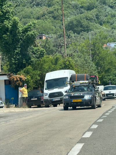 The image captures a sunny daytime scene on a paved road in Trap, Montenegro, with lush green hills and trees forming the backdrop. Several vehicles are parked or driving on the right side of the road, suggesting a traffic or parking area.
The primary focus is on a dark-colored van with a Dutch license plate reading "30-HN-LK". This van is loaded with equipment, including what appears to be a large gray bag secured to the roof rack with orange straps, and other items on its top, suggesting preparation for a journey or transport of goods.
In front of the dark van is a white van, also with a license plate visible that reads "BR-DJ952". Further down the road, another white vehicle is partially visible, with the license plate "BD-CC 723". Behind the dark van, another dark vehicle is parked, bearing the license plate "BD-AM580".
On the sidewalk next to the road, a man wearing a yellow t-shirt and a white cap is visible, appearing to be leaning against a railing or structure. His posture and the casual clothing suggest a relaxed atmosphere, possibly taking a break. The presence of houses and structures hints at a residential or mixed-use area.
The overall impression is that of a roadside scene in a somewhat rural or suburban area of Montenegro, with vehicles being a significant element, possibly indicating travel or local activity. The bright sunlight suggests clear weather.