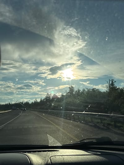 The image depicts a view from inside a vehicle driving on a highway. The sun is setting, casting a warm glow through the clouds and glinting off the windshield, which has several water spots. The highway curves to the left, with trees lining the right side of the road. Several vehicles are visible on the road ahead. The dashboard of the car is visible in the foreground. The scene appears to be in Krasnokamsk, Russia, given the context provided.
