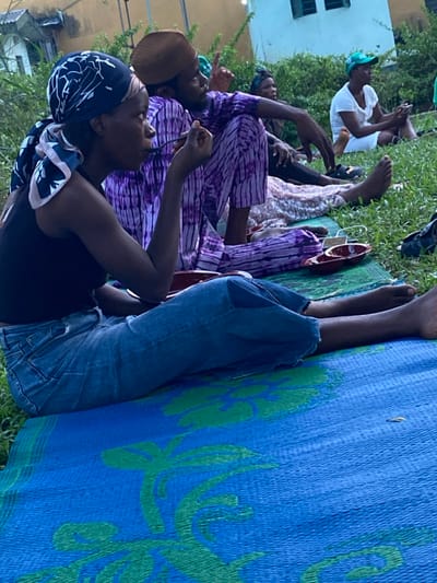 A group of people are seated on the grass and a patterned blue and green mat. Several people wear traditional purple and white clothing, while others wear casual attire like jeans and t-shirts. They appear relaxed, some interacting with their phones. In the background, a building with windows is visible, suggesting an outdoor gathering. The scene seems informal and communal.