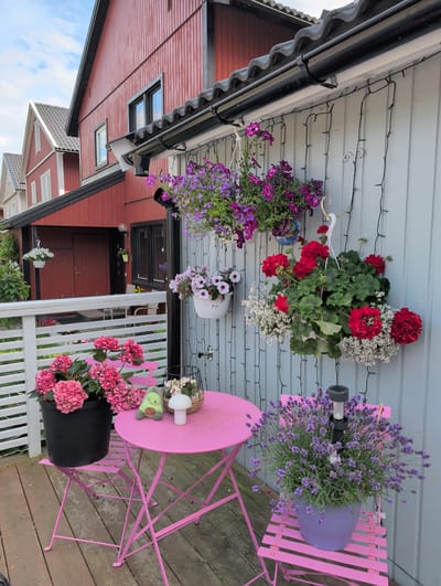A detailed, forensic, matter-of-fact description of the scene in Jordbro, Sweden:

The image captures a close-up view of a charming outdoor patio area, likely belonging to a residential property in Jordbro, Sweden. The foreground is dominated by a bright pink metal bistro set, consisting of a round table and two chairs, positioned on a weathered wooden deck. The table is laden with various decorative items: a black pot overflowing with vibrant pink hydrangeas, a small, light-green plush avocado toy, a white mushroom-shaped lamp, and a black wire basket holding a small cluster of white flowers. One of the pink chairs is occupied by a large purple pot filled with blooming lavender, topped with a solar-powered garden light.

The background reveals the exterior of a traditional Swedish house. The upper portion of the house is clad in vertical red wooden siding, with a dark grey corrugated roof. A black gutter system is visible along the edge of the roof. The lower section of the house, or an attached structure, features light grey horizontal siding. White string lights are draped vertically along the grey siding, adding a whimsical touch.

Several hanging flower baskets adorn the wall. One basket, to the left, contains a cascade of purple and pink petunias. Another, slightly to the right and higher up, holds red geraniums interspersed with smaller white flowers. Below these, a white hanging basket with white and pink flowers is also visible.

In the far background, the distinctive red architecture of neighboring houses is visible, suggesting a residential neighborhood. A white wooden fence runs horizontally across the middle ground of the image, separating the patio area from what appears to be a garden or yard beyond. The sky is partly cloudy. The scene conveys a sense of a well-maintained and decorated outdoor living space.