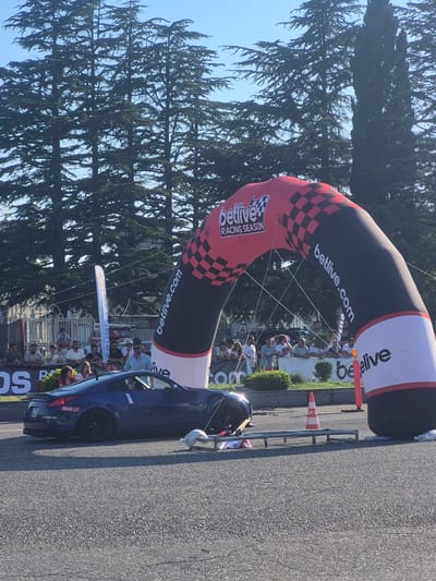 In the city of Poti, Georgia, a dark blue Nissan 350Z car is positioned on an asphalt road, appearing to be at the start or finish line of a race. The car is positioned under an inflatable archway that reads "betlive RACING SEASON" and "betlive.com". Spectators are gathered behind a barrier, observing the scene. The environment is outdoors with trees visible in the background and a clear blue sky overhead. There are orange traffic cones and a metal ramp-like structure on the road near the car.