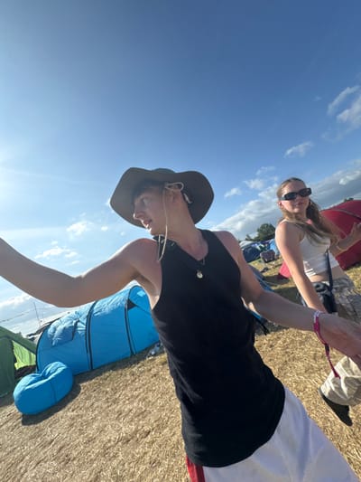 Two people pose against a partly cloudy sky. The person in the foreground is wearing a dark tank top, a hat, and light-colored bottoms. A blue tent is nearby. The second person, further in the background, sports sunglasses, a white top, and patterned shorts. They both appear to be outdoors during the day, possibly at a festival or event. The ground seems to be grassy.