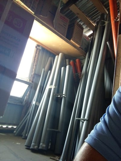 In a dimly lit storage space, possibly a garage or workshop, a collection of long, gray cylindrical objects, resembling pipes or conduits, are arranged vertically. Some of these objects have orange caps or accents. In the background, wooden shelving holds cardboard boxes, some with visible text and graphics. A window, partially obscured by a large cardboard box in the foreground, allows a bright light to enter the space. A person's arm, clad in a blue, textured shirt, is visible on the right side of the image, suggesting they are present in the scene. The environment appears cluttered and functional, indicative of storage or supply materials. The specific location within Kuybyshev, Russia, cannot be determined from the visual information provided in the image.