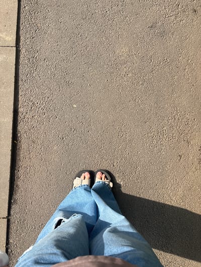 The image shows a pair of feet wearing light-colored sandals and blue jeans standing on an asphalt surface. A sidewalk is visible on the left side of the image. The sun is casting a long shadow to the right of the feet. The overall setting appears to be outdoors, possibly a street or pathway in a city.