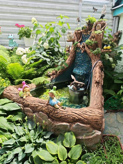 In Krutinskiy, Russia, a garden features a decorative pond with a log-shaped structure depicting a waterfall. The structure is adorned with various garden ornaments, including owls, squirrels, birds, and dragonflies. Two gnome figurines are placed on the log near the pond's edge. The surrounding area is filled with lush green foliage, including ferns, hydrangeas, and hostas, with some potted plants and a blue ceramic frog figurine also visible. The scene appears to be a tranquil outdoor setting.