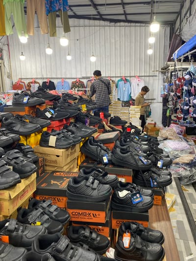 This image shows the interior of a shoe store in Lamteumen Barat, Indonesia. In the foreground, numerous pairs of black children's shoes are displayed on top of cardboard boxes. The shoes are mostly velcro-fastened sneakers, with some featuring visible brand labels like "COMEX" and "GENSTAR". Behind the shoes, more boxes are stacked, some with branding visible. In the mid-ground, several men are present. One man in a plaid shirt is standing and looking towards the right. Another man, wearing a light brown shirt, is looking down at his phone, standing near a display of clothing. Behind them, racks of clothing are visible, including shirts and pants in various colors. The store is illuminated by hanging lightbulbs, and the background features a corrugated metal wall. The overall scene depicts a busy retail environment with a focus on footwear and apparel.