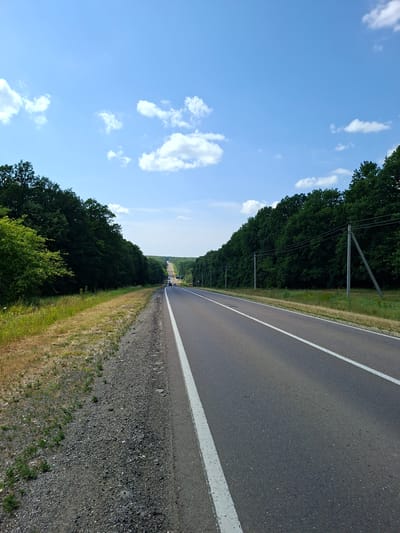 The image depicts a view of a paved road with a white line marking the lane, bordered by a gravel shoulder on the left and a dense line of trees on both sides. In the distance, the road curves and ascends a hill, with more trees visible on either side, eventually disappearing into the horizon under a clear blue sky with scattered white clouds. There are a few cars visible on the road in the distance. Utility poles with wires run along the right side of the road. The environment appears to be a rural or semi-rural area. The location is identified as Scoreni, Moldova.