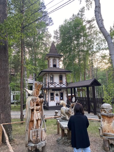 The image depicts an outdoor scene in the city of Tal'tsy, Russia, likely at an ethnographic museum or park. In the foreground, a wooden sculpture of a warrior in armor, complete with a crown, sword, and shield, stands prominently. To the right of this sculpture, a woman with dark hair, wearing a black jacket and light-colored pants, is standing, facing away from the camera and observing the scene. In the middle ground, a whimsical, multi-story building resembling a fairy-tale tower stands, with white walls, brick accents, and arched windows. A covered walkway extends from the building, leading to a dark wooden fence. Various other wooden sculptures, including a large cat-like creature and an owl-like bust, are scattered around the area. The background is dominated by tall, slender trees, characteristic of a forest or wooded park, with a bright, overcast sky overhead. The overall impression is of a rustic, fantastical, and culturally rich environment.