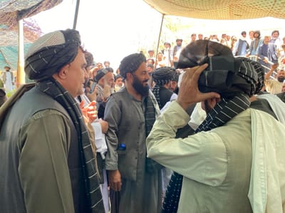 The image shows a group of men gathered under a white canopy, likely in the marketplace of Zaker-E Sharif, Afghanistan. The men are dressed in traditional Afghan clothing, including long tunics, trousers, and turbans. The man in the foreground, wearing a black and white turban, is adjusting his head covering with his right hand. Behind him is a man with a white turban who is looking off to the left side of the frame. Another man, wearing a black turban, is standing in the center of the image, with his back to the camera, adjusting his head covering. In the background, there is a crowd of people. The environment appears to be a bustling marketplace, with the sounds of people talking and haggling likely audible.  The presence of the white canopy suggests a temporary market structure or a gathering space for trading or socializing.