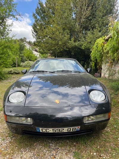A black Porsche 928 with a French license plate reading "CC-592-OB" is parked on a gravel driveway surrounded by grass. The car is facing the camera and is parked in front of a house. There is a large tree to the left of the car. The scene is located in Giverny, France, at coordinates 49.075, 1.532. The sky is blue with a few white clouds.