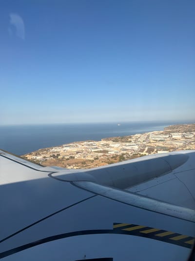 This image is an aerial view of the town of Birżebbuġa, Malta, as seen from an airplane window. The foreground is dominated by the wing of the aircraft, its smooth white surface punctuated by black and yellow hazard stripes and the text "NO STEP". Beyond the wing, the coastline of Birżebbuġa stretches along the Mediterranean Sea. The town appears as a dense collection of buildings with white roofs, interspersed with industrial structures and some green areas. The sea is a deep blue, and a faint haze blurs the distant horizon where a small boat is visible. The sky is clear and bright blue, with a subtle reflection of the airplane cabin visible in the upper left corner.
