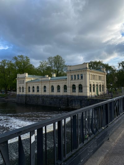 The image captures a partially obscured view of the Upplandsmuseet, a museum housed in an elegant, pale yellow building located on the banks of the Fyris River in Uppsala, Sweden. The building has classical architectural details, with arched windows and a detailed facade. It is partially obscured by trees in full leaf. To the right of the museum, a series of metal stairs descend into the flowing water of the river. The stairs are black and appear to be part of the riverbank's infrastructure. The sky above is overcast, with dark, heavy clouds dominating the frame, suggesting a potentially stormy weather condition. The angle of the photograph is from a high vantage point, potentially from a bridge or elevated pathway.