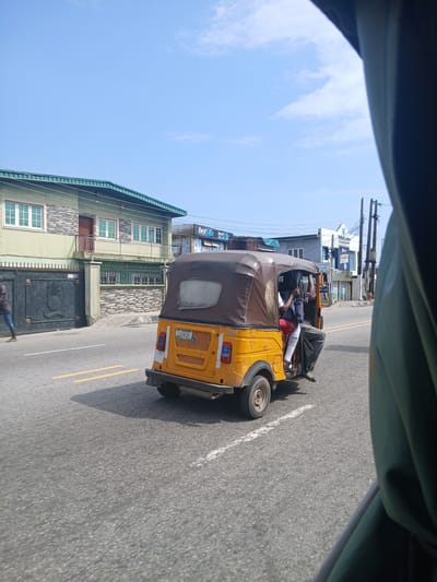 In Animashawun, Nigeria, on a bright, clear day, a yellow and brown auto-rickshaw, also known as a Keke Napep, is captured in motion. The vehicle is traveling on an asphalt road marked with yellow and white lines. The auto-rickshaw has a brown fabric roof and is carrying at least two passengers. One passenger is visible in the front, possibly the driver, wearing a white shirt and red shorts. Another passenger is seated behind, with their legs extending out of the vehicle. A third person, dressed in a dark t-shirt and blue jeans, walks on the side of the road in front of a large, dark metal gate. Behind the gate, a two-story building with a green roof and light green walls stands prominently. The building features multiple windows with white frames. To the right of the auto-rickshaw, a few shops with various signs, including one that reads "bet9ja" and another with "Vesali.com," are visible. Overhead, power lines and poles stretch across the sky, under a clear blue sky with scattered white clouds. The image appears to have been taken from inside another vehicle, as the edge of a dark green fabric is visible in the foreground.