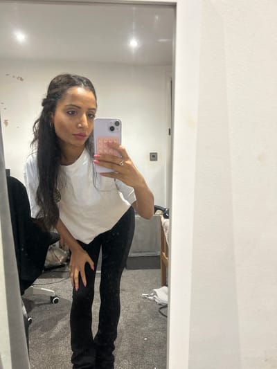 The image displays an adult female taking a mirror selfie in an indoor setting, identified as Winslow, United Kingdom. The subject, positioned centrally, is reflected in a full-length mirror, leaning forward with her left hand resting on her left thigh. She has long, dark, wavy hair, and is wearing a white short-sleeved t-shirt and dark-colored fitted pants. Her fingernails are painted pink, and she wears a ring on her left ring finger. She holds a light purple smartphone, equipped with a pink PopSockets-style accessory, in her right hand to capture the image.
The background visible in the mirror's reflection reveals a room with light-colored walls showing patches of discoloration or peeling paint on the upper left and right. A dark-colored square electrical switch or outlet is mounted on the wall to the right of her head. The floor is covered with a dark grey carpet, scattered with minor light-colored debris. Visible objects include a black office chair with white caster wheels on the far left, and a wooden furniture frame, possibly a bed frame or shelf unit, on the right, draped with a white textile. Two recessed ceiling lights are reflected, indicating overhead illumination. A white door frame is partially visible in the reflection towards the center-right.