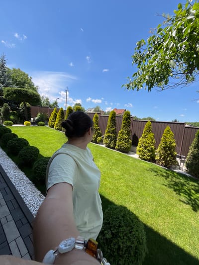This is a selfie taken outdoors on a sunny day, likely in the afternoon due to the long shadows cast across the lawn. The image features a person in the foreground, their back to the camera, with their dark hair styled in a bun. They are wearing a light yellow shirt and a beaded bracelet on their wrist.

The setting is a well-maintained backyard with a lush green lawn, manicured bushes, and a line of tall, cone-shaped evergreen trees. A tall, dark brown metal fence runs along the perimeter of the yard, behind the trees. In the distance, glimpses of houses and more trees are visible. The sky is a vibrant blue with scattered white clouds, indicating clear weather. The scene is one of relaxation and enjoyment of a pleasant outdoor environment.

There are no visible text elements in the image. The orientation of the photo is unusual, as if taken while the photographer was looking upwards or at an angle. The overall mood is peaceful and serene, capturing a moment of leisure in a suburban or rural Ukrainian landscape.