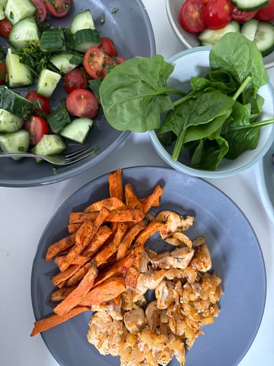 The image shows a close-up, overhead shot of a meal arranged on a white surface. There are three bowls and a plate visible. The bowl on the left contains diced cucumbers and halved cherry tomatoes, seasoned with herbs. The bowl above contains fresh spinach leaves. The plate to the right holds sweet potato fries and cooked chicken with onions. A fork rests in the bowl with the cucumber and tomato salad.
The image shows that the meal is a balanced combination of fresh vegetables, protein, and carbohydrates. There is nothing to imply the image was taken in Hazlemere, United Kingdom.