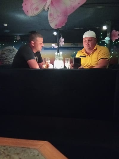Two men are seated at a dark table in what appears to be a dimly lit establishment. The man on the left, wearing a black t-shirt, is looking to his right with his mouth slightly open. In front of him is a glass containing a golden liquid, possibly beer. The man on the right, wearing a yellow polo shirt and a white skullcap, is looking down at a black smartphone he is holding. In front of him are two clear glasses, one appearing empty and the other with a small amount of liquid. The background is dark with decorative elements, including a large pink butterfly on the ceiling and pink flowers hanging from above. Some small white lights are visible in the background, suggesting a festive or decorative atmosphere. The overall scene suggests a casual meeting or outing.