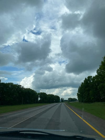 A wide-angle, eye-level, first-person perspective captures a view from a vehicle driving on a two-lane asphalt road. The road is lined with a bright yellow stripe on the right and a white stripe on the left, with lighter, striped markings on the road surface itself, possibly from recent rain. The road cuts through a rural landscape, with dense, dark green trees lining both sides of the road. The sky is filled with dramatic, grey and white cumulonimbus clouds, with patches of blue sky visible. The overall impression is that of driving through a slightly overcast, possibly humid, rural environment. No people or specific landmarks of Kushnarenkovo, Russia are identifiable.