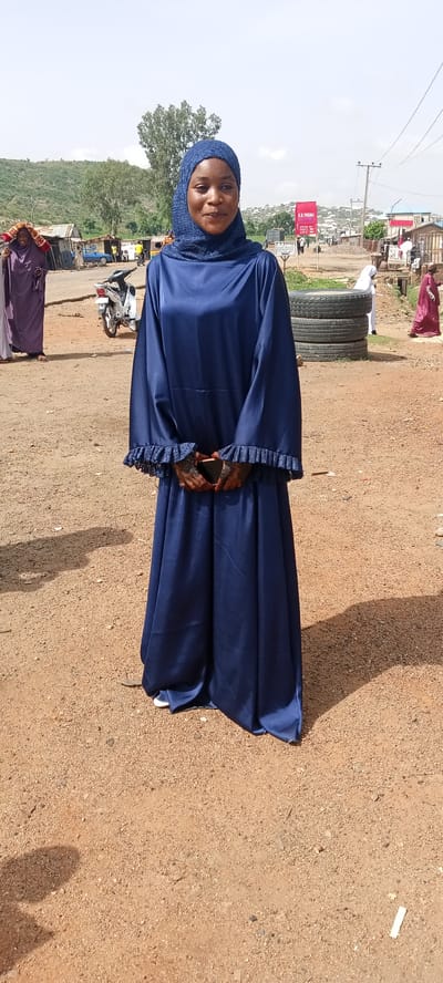 The image depicts a woman standing in what appears to be a street in Naraguta Hausawa, Nigeria. She is wearing a long, dark blue dress with lace trim at the wrists and a matching headscarf. Her hands are clasped in front of her, holding a phone.
In the background, there is a dirt road, various buildings, and a motorcycle. To the left, a person in a purple robe stands near the road. Behind the woman, there are stacked tires, and a sign with text is visible. Another person in a maroon robe is walking to the right. The landscape suggests a somewhat urbanized area with hills in the distance.