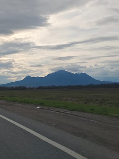 The image depicts a rural scene under a cloudy sky. A mountain range is visible in the distance, with some snow on the peaks. A field stretches between the viewer and the mountains, bordered by a line of trees. In the foreground, there is a road with a white line marking the edge. The road appears slightly wet or dirty. There is no indication of this location being Konstantinovskaya, Russia.