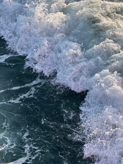 The image displays a close-up, dynamic view of turbulent seawater, positioned within the context of Palaiopoli, Greece. The scene is dominated by vigorous white foam and spray across the upper and right portions, indicative of breaking waves or agitated water. This foamy area exhibits varying densities, with countless individual water droplets visibly catching ambient light. The lower and left sections of the frame reveal the underlying darker blue-green water, characterized by undulating surfaces and patterns of swirling, dissipating foam trails. No individuals, vessels, or other man-made objects are present. The environment captured is purely aquatic, showing the natural interaction and movement of water under bright conditions.