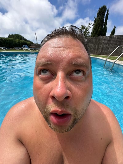 The image shows a close-up, fisheye perspective of a man's face partially submerged in a bright blue swimming pool. The man's hair is wet and slicked back, and his face is glistening with water droplets. His eyes are rolled upwards, and his mouth is slightly open, giving him a surprised or bewildered expression. The background features the pool's edge, a tiled bottom, a metal ladder, and lush green foliage and trees beyond a fence. The sky above is a bright blue with scattered white clouds. The scene appears to be outdoors on a sunny day, likely in a residential or resort setting within Sant Feliu de Guíxols, Spain.