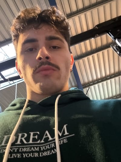 The scene depicts a young adult male in Chantepie, France, looking directly at the camera. The male has medium skin tone, dark curly hair, dark eyebrows, and a prominent mustache. A small hoop earring is visible in his left ear (viewer's left). He is wearing a dark green hooded sweatshirt with white drawstrings. White text on the sweatshirt reads, partially visible, "DREAM" followed by "DON'T DREAM YOUR LIFE / LIVE YOUR DREAM." The background reveals an indoor industrial-style environment featuring a light-colored corrugated metal ceiling and dark metal support beams. A dark metallic mechanism or device is partially visible in the upper right portion of the frame, attached to one of the beams. The image is captured from a low angle, looking upwards at the subject.
