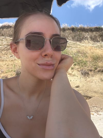 The image is a close-up, low-angle selfie taken outdoors on a sunny day. The primary subject is a young woman with light skin, wearing sunglasses and a silver necklace with a dragonfly pendant. Her face is partially obscured by her hand, which rests on her cheek. She appears relaxed and is looking at the camera.

The background is a dry, grassy hillside under a bright blue sky with scattered white clouds. The lighting suggests it is daytime, likely mid-morning or afternoon, given the clear sky and sunlight. The setting appears to be natural, possibly a park or a natural landscape, with hints of sand or light-colored rocks in the lower right corner. The woman is wearing a white top, possibly a swimsuit, indicated by the strap visible at the bottom of the frame. The image is cropped to focus on the woman's face, neck, and upper chest, with the background providing context for the outdoor setting. There is no visible text in the image.