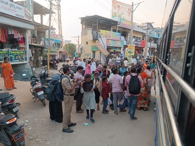 The image presents a busy street scene in Kothacheruvu, India. Approximately 25-30 individuals, mostly adults, are congregated near a blue and white bus that occupies the right side of the frame. The ground is an unpaved dirt surface with scattered debris.
Multi-story commercial buildings line the street. On the left, a building displays a sign for "Dr.P.V.SreeTeja BDS (Dental Surgeon)" and "దంత వైద్యశాల" (Dental Hospital), with clothing items visible for sale. A yellowish corner building in the mid-ground features "అన్నపూర్ణ హోటల్" and various food advertisements. Overhead utility poles, power lines, and a large cellular tower are visible against a bright, hazy sky.
Numerous motorcycles and scooters are parked along the street, with license plates such as "AP 02 RU 2790" and "AP 39R S7804" identifiable. An auto-rickshaw is partially visible further down the street.
Key individuals in the foreground include:
* A male police officer in a khaki uniform, black shoes, and a dark blue backpack, holding a device in his hands.
* A woman adjacent to the officer, wearing a black abaya, light blue hijab, and a dark blue backpack.
* A young girl in a light blue and white patterned dress, dark leggings, and blue sneakers, holding a dark bag.
* A boy in a maroon long-sleeved shirt and blue jeans.
* A woman in an orange saree is visible on the far left.
The crowd appears to be either waiting to board the bus or disembarking, with individuals standing and looking towards the bus or engaging in conversation.