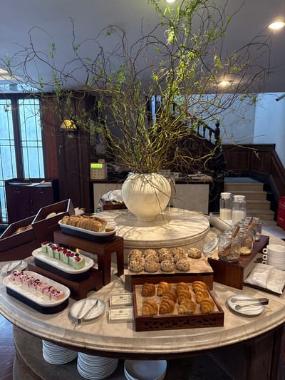The image depicts a well-lit indoor buffet scene, likely within a hotel or guesthouse dining area in Tongxiang, China. The focal point is a large, circular marble table laden with breakfast items. At the table's center, a substantial white ceramic vase holds an arrangement of bare and budding branches, serving as a decorative centerpiece.

On the table, various food items are displayed on wooden risers and platters. To the left, two platters feature rectangular desserts: the lower one containing pink, layered treats, and the upper one presenting green, layered desserts garnished with berries. A large loaf of bread rests on a white dish on a taller wooden stand. Further to the right, a selection of round bread rolls, many topped with seeds, are arranged on a wooden tray. Below these, a tray of golden-brown croissants is visible. Two small white signs with Chinese text accompany the pastries.

Adjacent to the food, serving implements such as metal tongs are positioned next to empty plates stacked vertically. On the right side of the table, clear glass bottles containing milk and multiple glass jars filled with cereals or granolas, each with a wooden spoon, are arranged on another wooden riser. A stack of white cups or bowls is present, along with a few loose cinnamon sticks.

In the background, dark wooden paneling and furniture are visible. To the far left, a window with vertical bars allows natural light into the space. A dark wooden cabinet with a serving tray is positioned beneath this window. Towards the center-left background, a cream-colored electronic device with multiple yellow indicator lights is visible. To the right, a dark wooden staircase with a prominent banister ascends, suggesting a multi-level interior. The overall environment conveys a refined, established atmosphere. No individuals are present in the image.