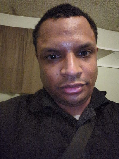 A close-up, slightly blurry, headshot of a dark-skinned man. His head is slightly tilted to the right. He has short, dark hair. He's wearing a dark shirt with a gray collar. The background appears to be a beige wall with white molding. The lighting is somewhat uneven. His face is the clear focus of the image.