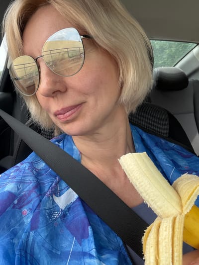 A woman with short blonde hair, wearing large, round sunglasses and a blue, abstract-patterned shirt, is seated in the passenger seat of a car. She is holding a partially peeled banana in her right hand, bringing it towards her mouth. Her left hand is not visible. A black seatbelt is visible across her chest. The background of the car interior, including a dark headrest and a window showing greenery outside, is visible. The image does not contain any information about the city of Teshebs, Russia.