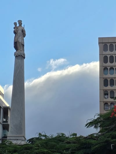 The image depicts a monument topped with a statue of a woman holding a child, likely a religious figure, in Santa Cruz de Tenerife, Spain. The monument is made of light-colored stone and is an obelisk shape. To the right of the monument, a modern building with distinctive oval-shaped windows is visible. In the foreground, lush green foliage with some red flowers is present. The sky above is a mix of blue and white clouds.