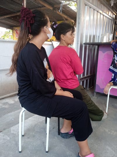 The image shows three people sitting on chairs in an outdoor setting. In the foreground, a person with long, dark, wavy hair pulled back in a high ponytail with a burgundy accessory, wearing a black long-sleeved shirt and black pants, sits on a white stool. This person is wearing a white face mask and dark clogs with pink accents. To their right, another person with dark hair pulled back, wearing a bright red t-shirt and olive green pants, sits on a dark stool, facing away from the camera and looking to the right. Further to the right, a person in a blue shirt with white and black accents is partially visible, with their bare foot resting on a white stool. They appear to be a child. The background consists of a corrugated metal structure, possibly part of a stall or building, with some wooden beams and roofing overhead. There are hints of vegetation and other structures in the distance. The ground is paved with concrete. The scene suggests a casual outdoor gathering or waiting area, possibly in a market or roadside establishment in Punden, Indonesia.