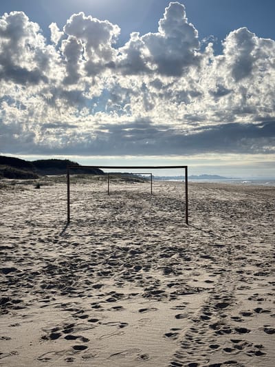 The image depicts a beach in Ciudad de la Costa, Uruguay. The foreground is sandy beach, marked with numerous footprints and ripple patterns. Four evenly spaced, dark-colored rectangular frames stand upright, running parallel to the shoreline. Beyond these frames, there is a line of vegetation that slopes upward. In the distance, a faint horizon line is visible where the sea meets the sky. To the left, large, billowing white clouds fill the sky, contrasting against a blue backdrop. The sun is faintly visible behind the clouds, creating a bright glow.