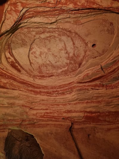 The image shows a close-up of a sandstone formation in the Schlossberghöhlen caves of Homburg, Germany. The rock exhibits a swirling, concentric pattern, with layers of red and beige hues. A small, round hole is visible in the upper left of the concentric pattern. Cracks and fissures are present throughout the rock surface. In the bottom right, a darker area suggests a deeper part of the cave structure.