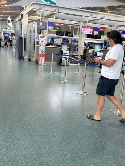 A man with dark, wavy hair, wearing a white t-shirt and navy blue shorts, walks through an airport terminal in Tambon Mai Khao, Thailand. He is looking at his smartphone, which he holds in his right hand. The man is wearing dark sandals. The terminal has a green tiled floor and several check-in counters with digital displays. Some displays show "AirAsia" and flight information. Metal stanchions with blue ropes cordon off waiting areas. In the background, a large white triangle with a green stripe, labeled "B", is visible, indicating a section of the terminal. The overall environment suggests a busy airport setting.